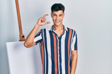 Genç İspanyol bir adam boş tuvalin yanında durup gülümsüyor ve el hareketleriyle küçük boy işareti yapıyor ve kameraya bakıyor. ölçüm kavramı. 