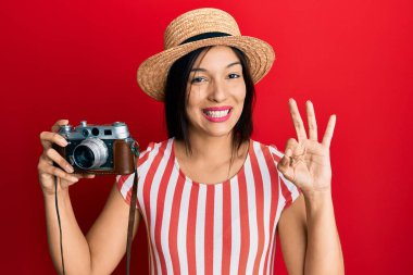 Yazlık şapka takan genç Latin bir kadın elinde klasik kamerayla tamam işareti yapıyor, gülümseyerek mükemmel bir sembol oluşturuyor. 