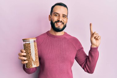 Sakallı genç adam elinde sağlıklı tahıl gevreği kavanozuyla gülümsüyor bir fikir veya soru işareti ile mutlu bir yüzle, bir numara 