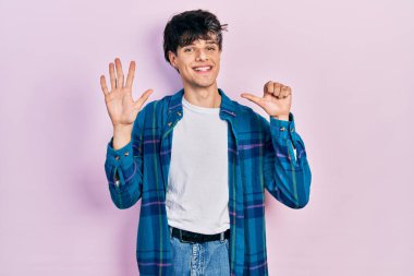 Yakışıklı hippi genç adam gündelik beyaz tişört ve klasik tişört giyiyor. Altıncı parmaklarını gösterip gösterirken kendinden emin ve mutlu gülümsüyor.. 