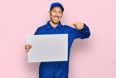 Sakallı, inşaatçı tulumu giymiş, elinde boş pankart tutan kel bir adam mutlu ve gururlu bir kendini işaret ediyor. 