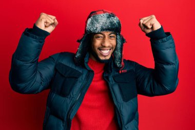 Sakallı, kışlık şapkalı ve paltolu genç Afrikalı Amerikalı adam kollarını kaldırarak ve açık gözlerle başarıyı kutluyor ve şaşırıyor. Kazanan konsept. 