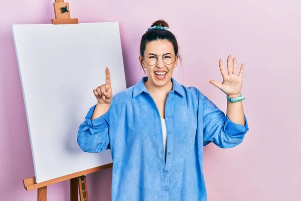 自信と幸せ笑顔ながら 画家のイーゼルスタンドに立っている若いヒスパニック系の女性は指の数を表示し 指摘します — ストック写真