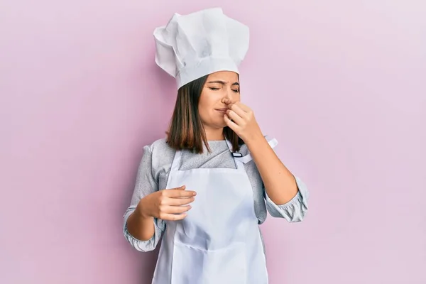 年轻美丽的女人穿着职业厨师制服 头戴帽子 闻到一股难闻难闻的气味 用手指捂住鼻子呼吸 — 图库照片