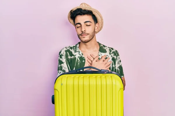 Young hispanic man wearing summer style and holding cabin bag smiling with hands on chest, eyes closed with grateful gesture on face. health concept.