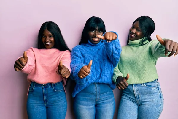 Three young african american friends wearing wool winter sweater approving doing positive gesture with hand, thumbs up smiling and happy for success. winner gesture.