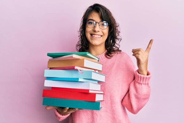Young hispanic woman holding a pile of books smiling happy pointing with hand and finger to the side 