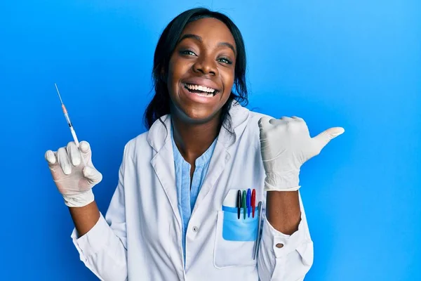 Joven Mujer Afroamericana Vistiendo Uniforme Científico Sosteniendo Jeringa Apuntando Pulgar —  Fotos de Stock