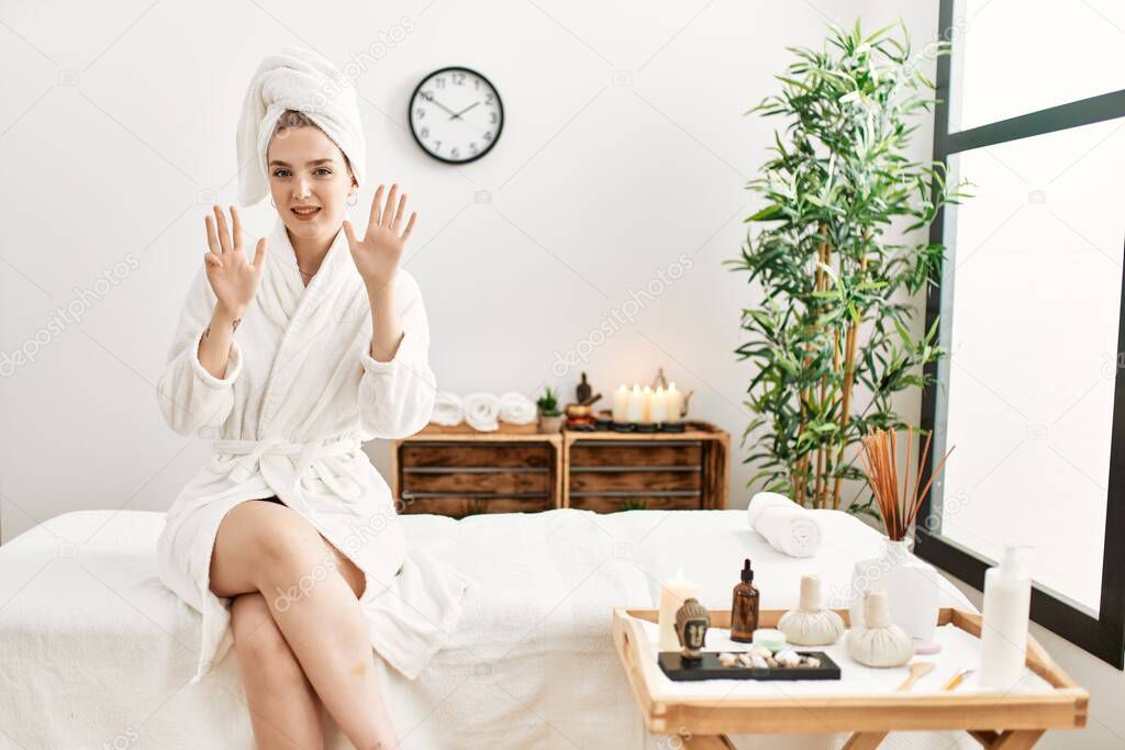 Young blonde woman wearing bathrobe at wellbeing spa showing and pointing up with fingers number nine while smiling confident and happy. 
