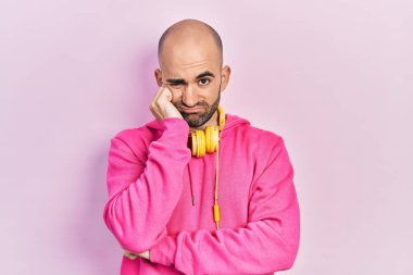 Spor kıyafetleri giyen ve kulaklık takan genç kel bir adam kollarını kavuşturarak depresyondan sıkılmış ve yorgun göründüğünü düşünüyor.. 