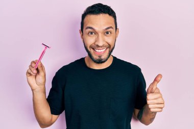 Sakallı İspanyol adam elinde jilet, mutlu ve pozitif gülümsüyor, başparmağıyla mükemmel ve onay işareti yapıyor. 