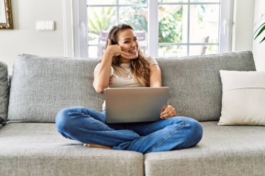 Güzel, genç, esmer bir kadın koltukta oturup bilgisayarını kullanarak gülümsüyor telefonla el ve parmaklarıyla telefonla konuşuyor gibi yapıyor. iletişim kavramları. 