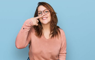 Genç ve cüsseli bir kadın, günlük kıyafetler giyiyor ve gözlük takıyor. Parmaklarıyla ve burnuyla işaret ediyor. Neşeli gülümsüyor. güzellik konsepti 