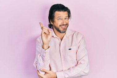 Orta yaşlı, günlük kıyafetler giyen, yüzünde kocaman bir gülümseme olan beyaz bir adam, kameraya el ve parmak ile bakıyor.. 