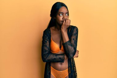 Genç Afro-Amerikalı kadın iç çamaşırı giyiyor stresli ve gergin görünüyor elleri ağızda, tırnaklarını yiyor. anksiyete. 