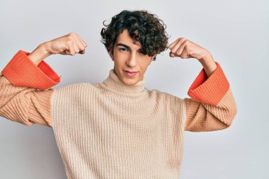 Sıradan bir kazak giyen İspanyol genç adam gururla gülümseyen kol kaslarını gösteriyor. fitness konsepti. 