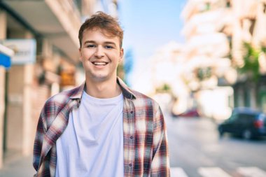 Genç beyaz adam gülümseyerek şehirde mutlu mesut duruyor..