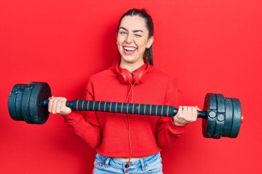Mavi gözlü güzel bir kadın spor kıyafetleri giyiyor, dambıllar kullanıyor, kameraya seksi bir ifadeyle bakıyor, neşeli ve mutlu bir yüzle.. 