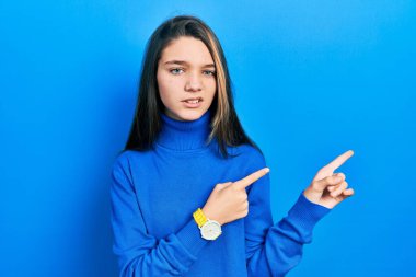 Balıkçı yaka kazak giyen genç esmer kız iki eliyle endişeli ve endişeli bir ifadeyi işaret ediyor. 