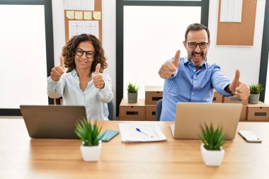 Orta yaşlı İspanyol kadın ve erkek ofiste dizüstü bilgisayarlarıyla oturup elle olumlu jest yapmayı onaylıyor, gülümsüyor ve başarı için seviniyor. kazanan jest. 