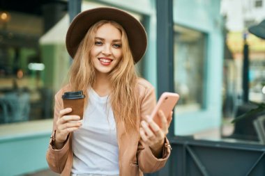 Genç sarışın kız akıllı telefon kullanıyor ve şehirde kahve içiyor..