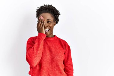 Genç Afrikalı Amerikalı kadın, izole edilmiş arka planda günlük giysiler giyiyor. Bir gözünü eliyle kapatıyor. Yüzünde kendine güvenen bir gülümseme ve sürpriz bir duygu var.. 