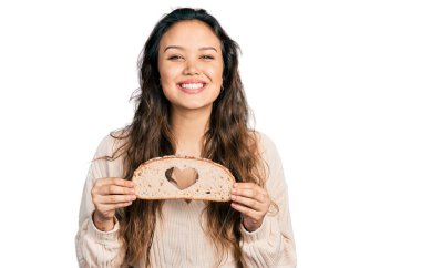 Genç İspanyol kız elinde kalp simgesi olan ekmek tutarken yüzünde mutlu ve havalı bir gülümsemeyle gülümsüyor. diş gösterme. 