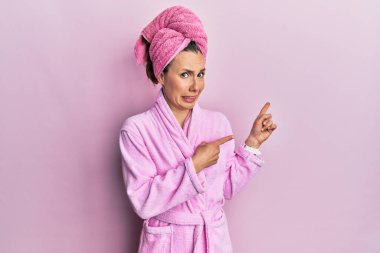 Duş başlığı ve bornoz takan genç sarışın kadın endişeli ve gergin bir şekilde iki elini de işaret ediyor. Endişeli ve şaşırmış bir ifade. 