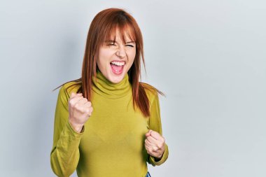 Balıkçı yaka kazak giyen kızıl saçlı genç kadın kolları havada ve gözleri kapalı bir şekilde başarıyı kutluyor. 