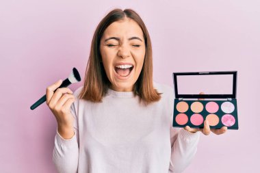 Makyaj fırçası tutan genç ve güzel bir kadın çılgınca kutlama yapıyor ve şaşkınlık içinde gözleri açık heyecanla bağırıyor.. 