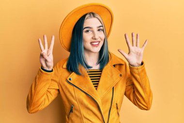 Sarı şapkalı ve deri ceketli genç modern bir kız sekiz numaralı parmaklarıyla gülümseyerek mutlu ve kendinden emin bir şekilde onu işaret ediyor.. 