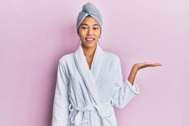Duş havlusu şapkası ve bornoz takan genç Çinli kadın neşeli bir şekilde gülümsüyor ve avuç içi kameraya bakıyor.. 
