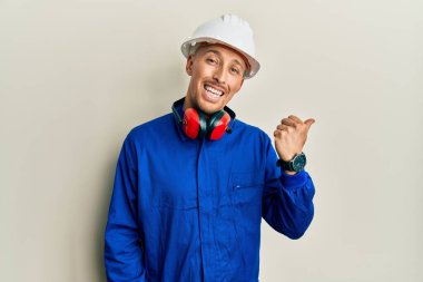 Sakallı, inşaatçı üniforması giyen, kasketli, gülen yüzlü ve baş parmağıyla yüzünü gösteren kel bir adam.. 