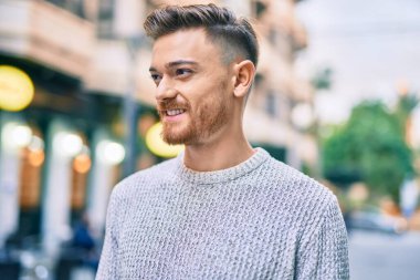 Genç beyaz adam gülümseyerek şehirde mutlu mesut duruyor..