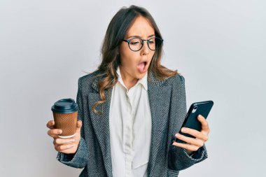 Genç esmer kadın akıllı telefon kullanıyor ve bir fincan kahve içiyor şaşkınlık ve şaşkınlık içinde ifade, korku ve heyecanlı yüz ifadesiyle.. 