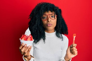 Afro saçlı Afrikalı bir kadın çilekli dondurma yiyor şüpheci ve gergin, sorun yüzünden surat asıyor. negatif kişi. 