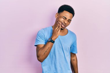Sıradan mavi tişörtler giyen genç Afro-Amerikan bir adam diş ağrısı ya da diş hastalığından dolayı acı dolu bir ifadeyle dudaklarına dokunuyor. dişçi 