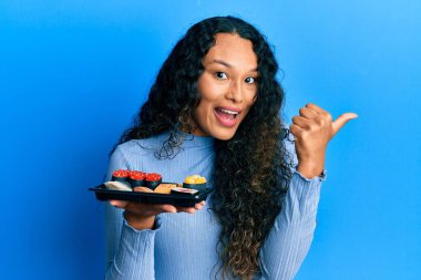 Suşi tabağını gösteren genç Latin bir kadın başparmağıyla yan tarafa bakıyor ve ağzı açık gülümsüyor. 