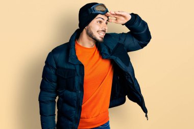 Kar maskesi ve gökyüzü gözlüğü takan, sakallı, mutlu ve tebessüm eden genç bir İspanyol. arama kavramı. 