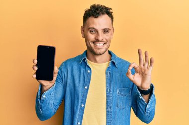 Genç İspanyol adam elinde akıllı telefonuyla ekranda boş görünüyor. Parmaklarıyla işaret veriyor, gülümsüyor. Mükemmel bir sembol. 