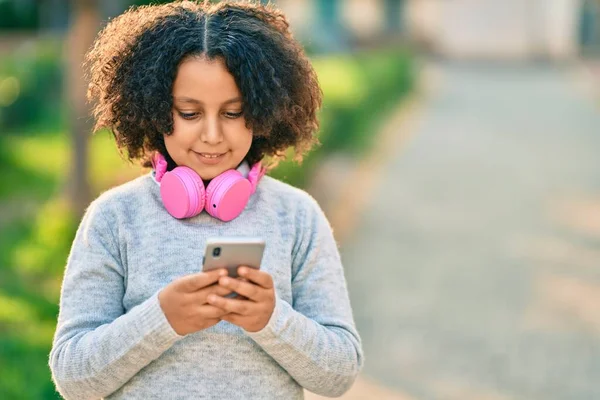 公園でスマートフォンやヘッドフォンを使用して音楽を聴く愛らしいヒスパニック系の子供の女の子 — ストック写真