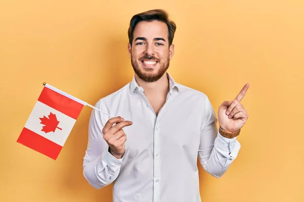 英俊的高加索人 留着胡子 拿着加拿大国旗 高兴地微笑着 手指手画脚地指向旁边 — 图库照片