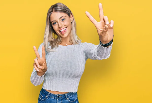 Beautiful blonde woman wearing casual clothes smiling with tongue out showing fingers of both hands doing victory sign. number two.