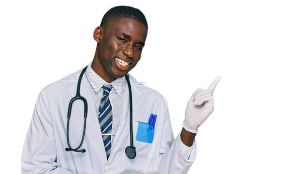 穿着医生制服和听诊器的年轻的非洲裔美国人高兴地微笑着 手指头指向旁边 — 图库照片