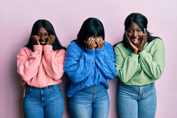 Three young african american friends wearing wool winter sweater laughing and embarrassed giggle covering mouth with hands, gossip and scandal concept