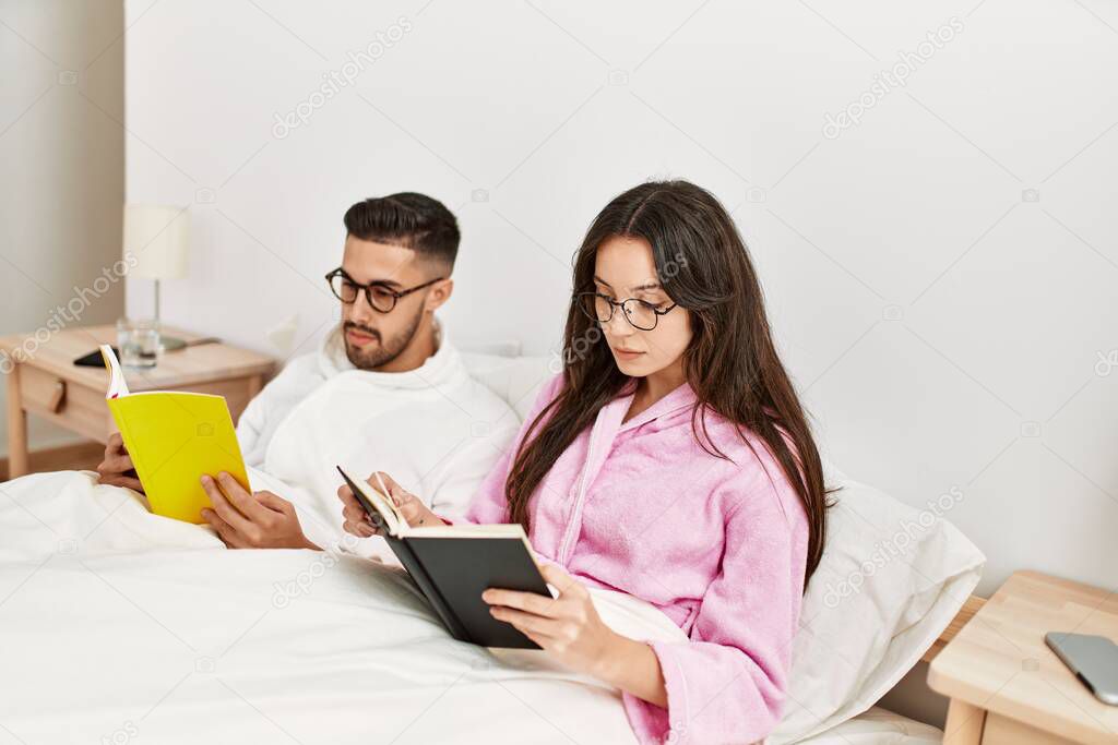 Young hispanic couple reading book lying in bed at home.