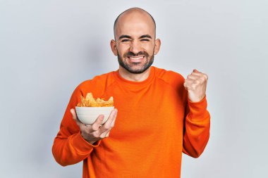 Genç kel adam patates cipsi tutuyor. Gururla bağırıyor, zaferi kutluyor ve başarıyı kollarını kaldırarak kutluyor. 