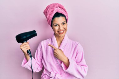 Duşta bornoz giyen, kurutma makinesi kullanan, gülümseyen, mutlu, eli ve parmağı olan genç İspanyol kadın. 