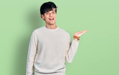 Yakışıklı hippi genç adam sıradan bir kazak giyiyor neşeli bir şekilde gülümsüyor ve avuç içi kameraya bakıyor.. 