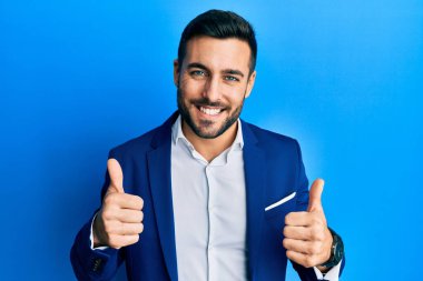Genç İspanyol iş adamı, iş ceketi giyerek olumlu jest yapıyor, gülümsüyor ve mutlu. Neşeli ifade ve kazanan jest. 